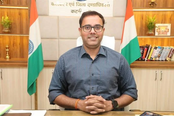 कार्यालय में मेज के पीछे बैठे चश्मा पहने अधिकारी, दोनों ओर भारतीय ध्वज दिखाई दे रहे हैं